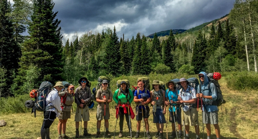 A group of students wearing backpacks pose for a photo in a green meadow